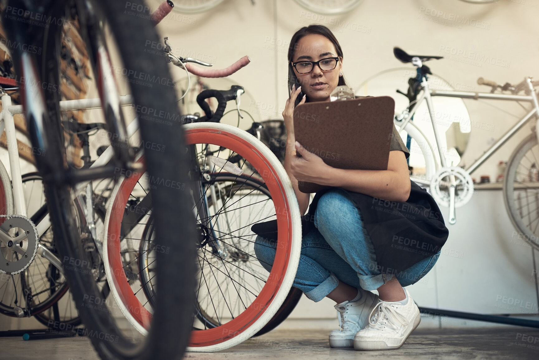 Buy stock photo Full length shot of an attractive young woman crouching in her bicycle shop and using her cellphone while holding a clipboard