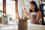 Paintbrush, art and equipment with a jar on a table in a pottery workshop or studio and a designer woman in the background. Zoom, paint and creative with a female potter working on a design