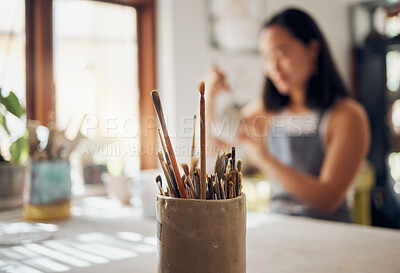 Buy stock photo Paintbrush, art and equipment with a jar on a table in a pottery workshop or studio and a designer woman in the background. Zoom, paint and creative with a female potter working on a design