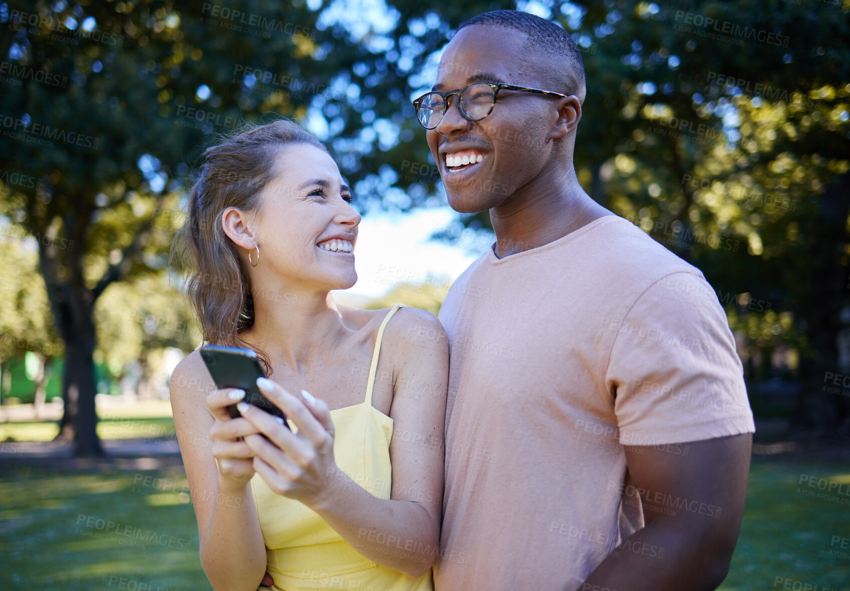 Buy stock photo Comedy, laughing and interracial couple with a phone in nature, funny communication and smile at a meme. Comic, streaming and black man and woman reading a joke on a mobile in a park in France