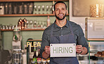 Small business, man or business owner with a hiring sign for job vacancy offer in cafe or coffee shop. Boss, marketing or happy entrepreneur smiles with an onborading recruitment message in store
