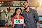 Portrait, small business or couple with open sign to welcome sales in a cafe or coffee shop with hospitality.  Restaurant or managers with a happy smile with message on board after opening a store