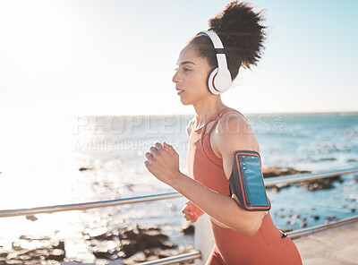 Buy stock photo Black woman, fitness and running with headphones and cellphone on arm at the beach in Cape Town. Sporty African American female runner by the ocean coast having a run for cardio training workout