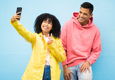 Buy stock photo Friends, phone and peace sign for selfie on a blue background for fashion, style or friendship together. Young man and woman smiling and looking at smartphone for photo leaning on a wall in happiness