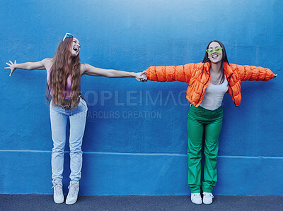 Buy stock photo Friends, wall and city for fashion with girl, holding hands and outdoor for funny, comic face or happy together. Gen z women, metro and blue background for trendy clothes, sneakers or urban adventure
