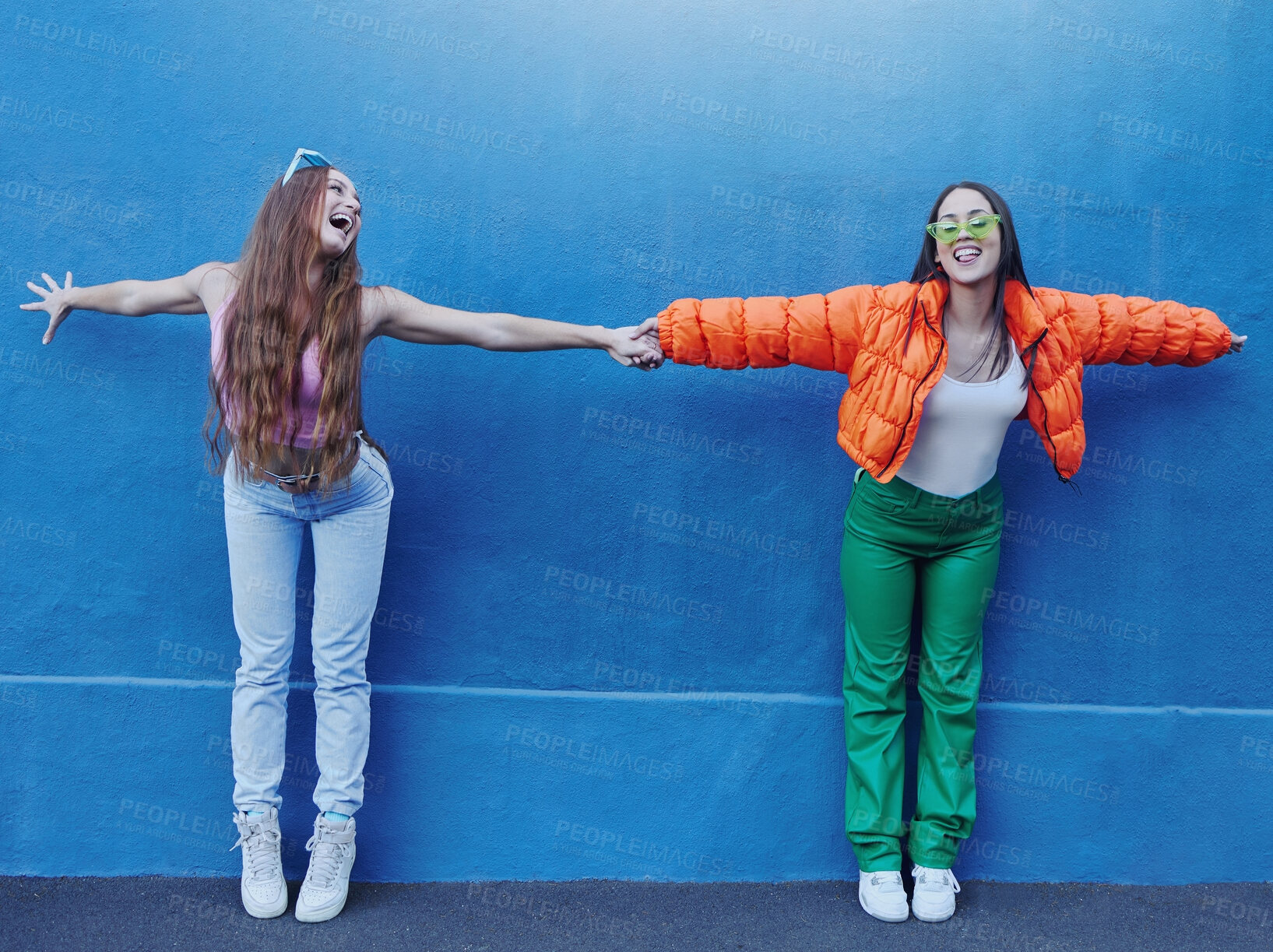 Buy stock photo Friends, wall and city for fashion with girl, holding hands and outdoor for funny, comic face or happy together. Gen z women, metro and blue background for trendy clothes, sneakers or urban adventure
