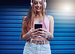 Social media, music and woman reading on a phone with a podcast, communication and app in the city. Headphones, radio and girl typing on a mobile, streaming audio and sound on a blue wall in Germany