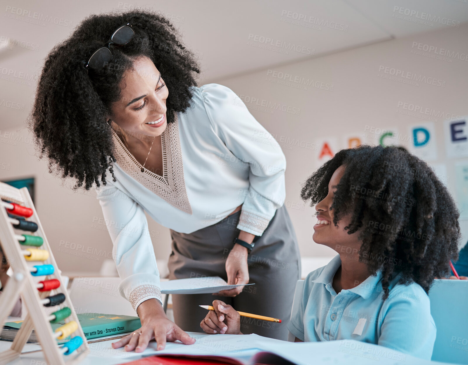 Buy stock photo Female teacher in classroom, helping girl student assessment and writing notes in book with pen. Learning math together, educator reading notebook and listening to child answer question at school