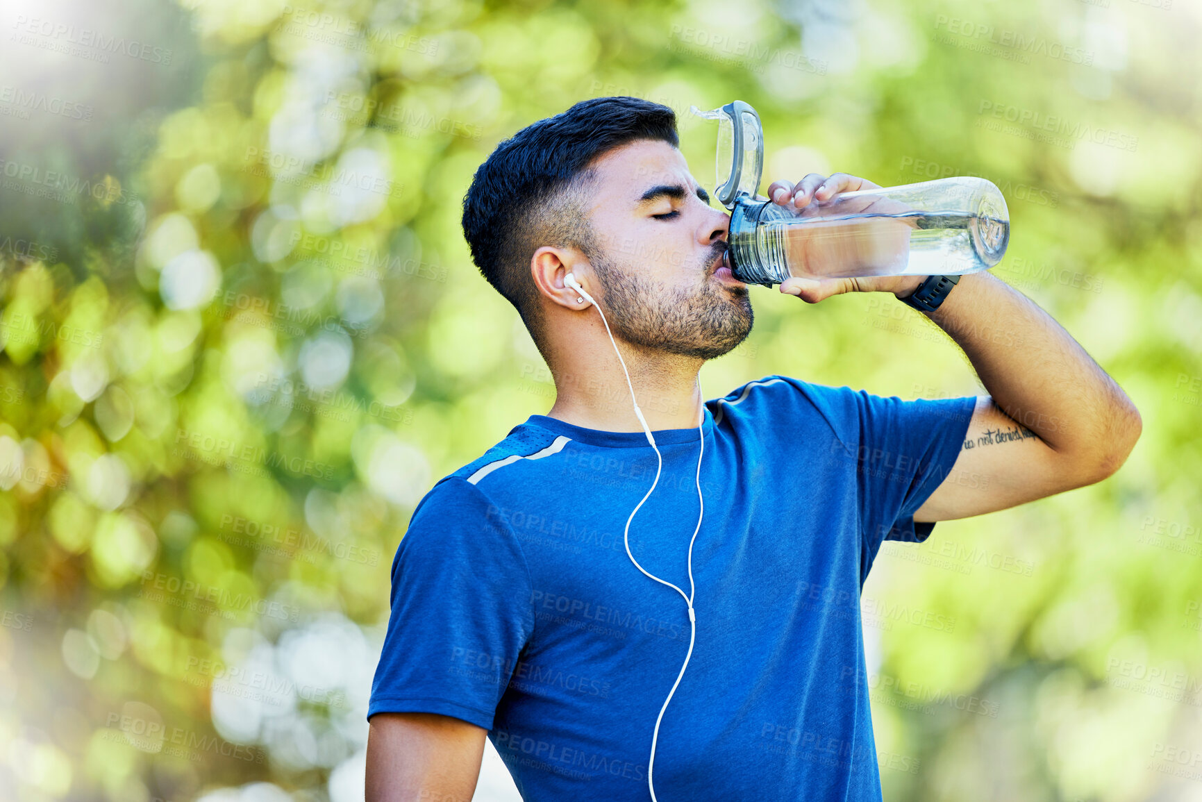 Buy stock photo Nature, running and thirst, a man drinking water on fitness break with headphones in park. Health, exercise and freedom, runner in forest with a drink from bottle while on run in natural landscape.