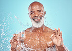 Splash, portrait and elderly man in a studio with a glass of water and a ok hand gesture. Health, wellness and happy senior African male with sign language by a blue background with mockup space