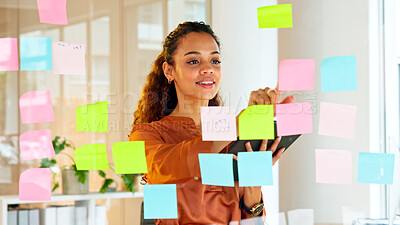 Happy female designer planning ideas on a glass wall with colorful sticky notes inside a creative and modern office. Busy woman enjoying her job while brainstorming projects and managing projects