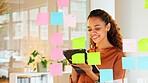 Happy female designer planning ideas on a glass wall with colorful sticky notes inside a creative and modern office. Busy woman enjoying her job while brainstorming projects and managing projects