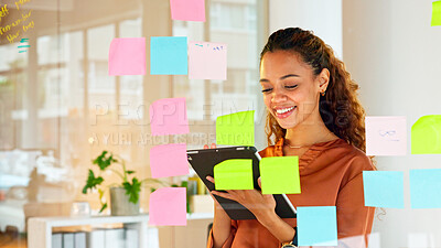 Happy female designer planning ideas on a glass wall with colorful sticky notes inside a creative and modern office. Busy woman enjoying her job while brainstorming projects and managing projects