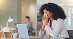 Sick businesswoman blowing nose with a tissue and suffering from flu virus or sinuses while working on a laptop in a modern office. Professional female with afro feeling ill while busy typing email