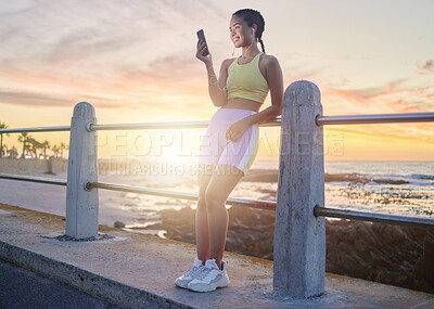 Buy stock photo Fitness, beach and woman with smartphone after running at sunset, online and streaming music or podcast in nature for exercise. Runner, relax and scroll social media after run, smile with earphones.
