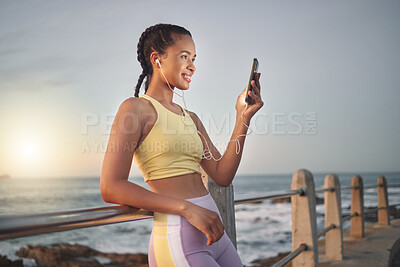 Buy stock photo Fitness, beach and woman with smartphone after running at sunset, online and streaming music or podcast in nature for exercise. Runner, relax and scroll social media after run, smile with earphones.