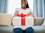 Christmas, gift and a box in the hands of a black woman in her home, sitting on the living room sofa. Birthday, present and event with a female giving a package during a celebration surprise