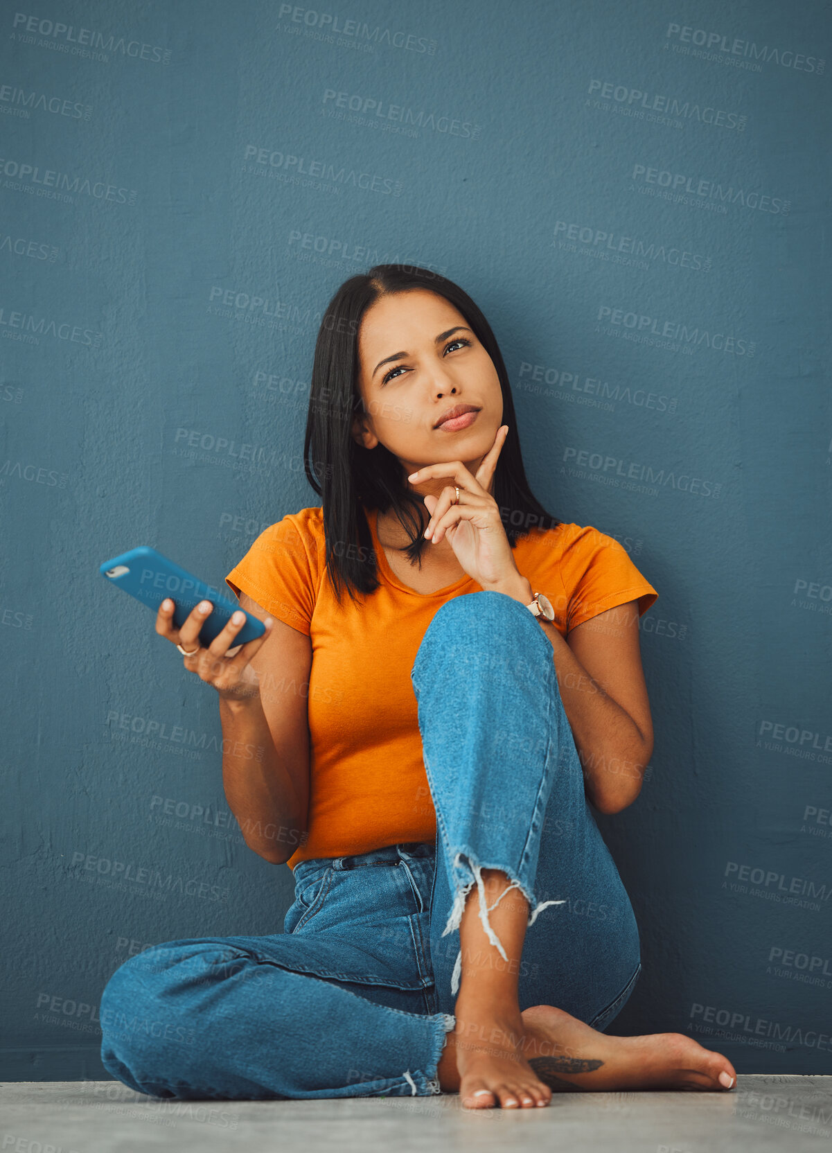 Buy stock photo Phone, thinking and social media with a woman in studio on a blue background, sitting on the floor. Mobile, contact or idea and an attractive young female posing with her finger on her chin