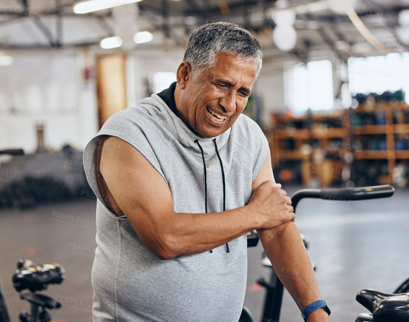 Buy stock photo Sports, gym and injury, old man with arm pain, emergency during workout at fitness studio. Health, wellness and inflammation, senior person with hand on muscle cramps while training on exercise bike.