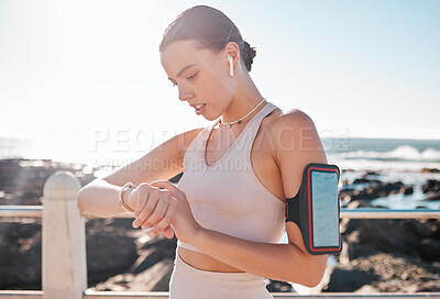 Buy stock photo Fitness time, smartwatch and woman with headphones checking running goals by the sea. Ocean, exercise app and workout timer with a runner by ocean water in summer after run and sports by a beach