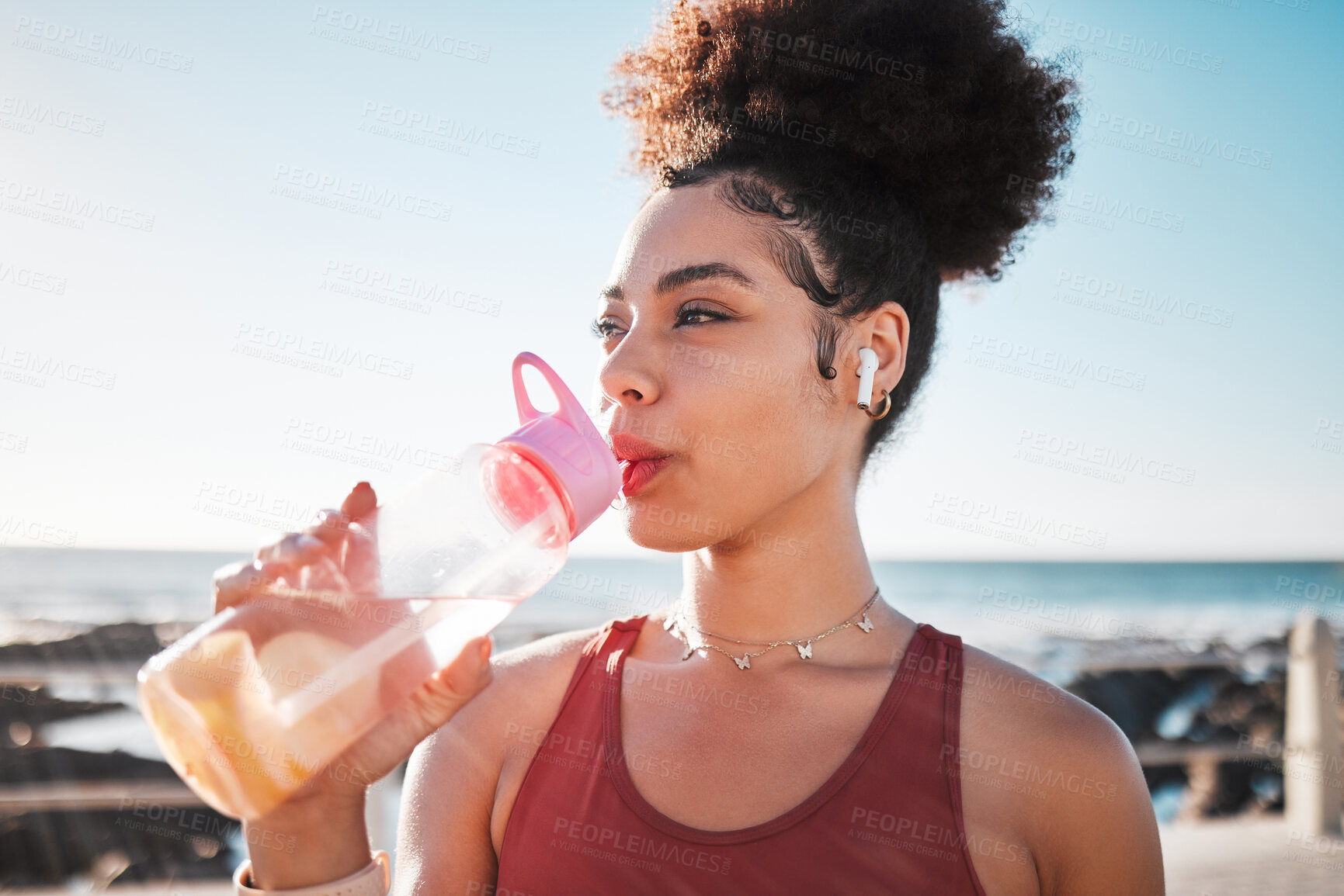 Buy stock photo Fitness black woman drinking water on beach for exercise, listening to music and cardio training in blue sky lens flare.  Liquid bottle for diet, goals and tired sports runner or USA person by ocean