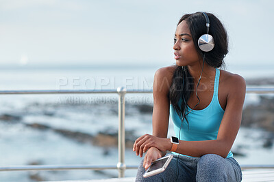 Buy stock photo Black woman, phone and headphones at ocean, sitting and thinking with idea, music and relax to focus. Runner, rest and smartphone with vision, social media app and mindfulness on outdoor adventure