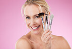 Portrait, makeup and brushes with a mature woman in studio on a pink background to promote a cosmetic product. Face, happy and cosmetics with an attractive older female posing for beauty treatment