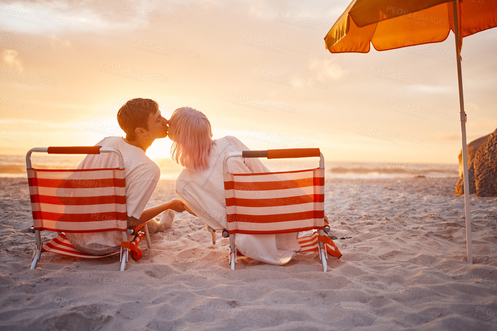 Buy stock photo Couple, kiss and beach chairs by a ocean sunset with love, happiness and care on vacation. Sea, sunshine and kissing of happy young people together sitting by the sand feeling relax outdoor 