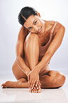 Portrait, beauty and body with a model black woman in studio sitting on the floor against a gray background. Health, wellness and skincare with an attractive young female touching her foot inside