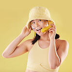 Woman, banana phone call and studio with smile, communication and eyes sticker art on face. Asian model girl, yellow background and fashion with beauty, aesthetic and cosmetic wellness with fruit