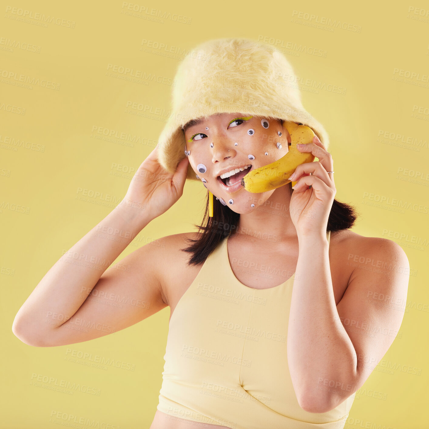 Buy stock photo Woman, banana phone call and studio with smile, communication and eyes sticker art on face. Asian model girl, yellow background and fashion with beauty, aesthetic and cosmetic wellness with fruit