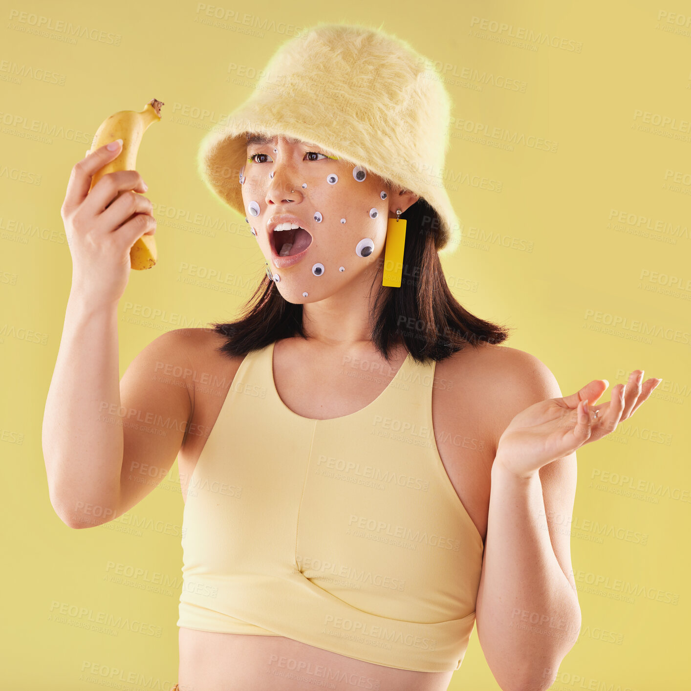 Buy stock photo Shock, surprise and woman with a banana in a studio with creative eye stickers on her face. Fruit, fashion and Asian female model with omg, wtf or wow facial expression isolated by yellow background.