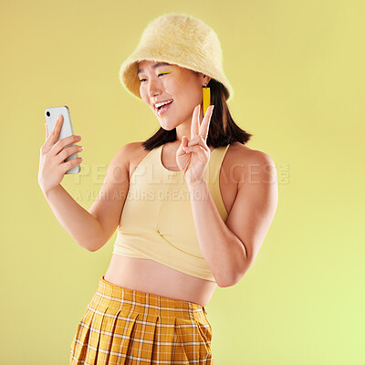 Buy stock photo Selfie, peace sign and smile of Asian woman in studio isolated on a yellow background. Makeup, fashion hat and young female model with v hand gesture taking pictures for social media or happy memory.