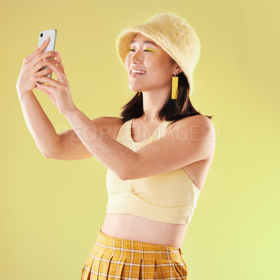 Selfie, beauty and smile of Asian woman in studio isolated on a yellow background. Makeup, fashion hat or young female model taking pictures or photo for social media, profile picture or happy memory