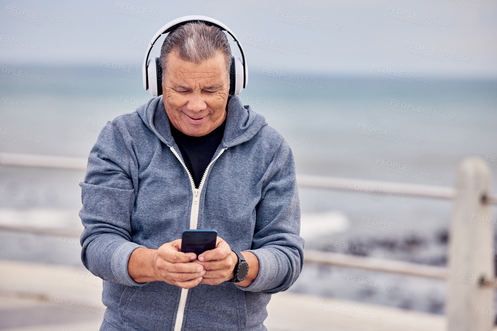 Buy stock photo Music, phone and senior man by ocean for fitness, exercise and training for healthy lifestyle on promenade. Retirement, headphones and elderly male listening to audio, track or radio and texting