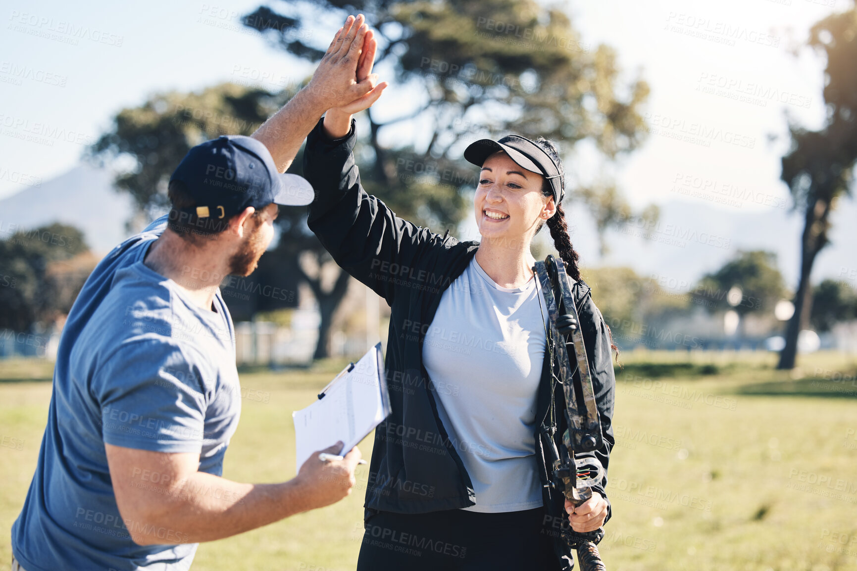 Buy stock photo Woman, archery and personal trainer high five in sports for partnership, success or celebration on the grass field. Happy sporty female smile and touching hand of male coach for archer sport training