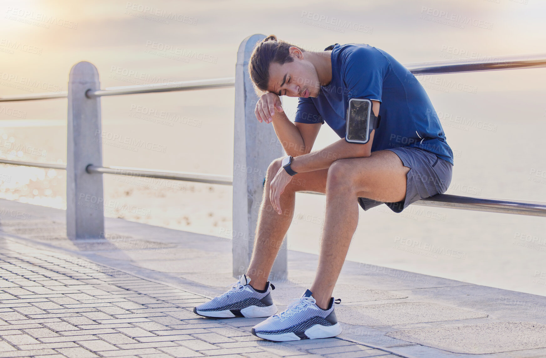 Buy stock photo Tired runner man, sitting and beach for workout, health and exercise with headache, rest and relax. Gen z guy, training burnout and challenge with breathing, mindset and fitness on railing by ocean