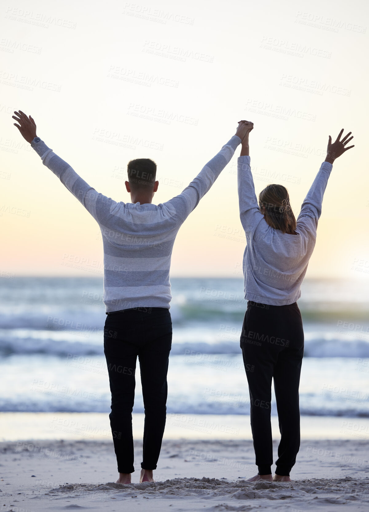 Buy stock photo Couple, beach and hands up outdoor while happy at sunset for love, freedom and peace with calm ocean. Young man and woman together on vacation holiday at sea to relax, travel and connect in nature