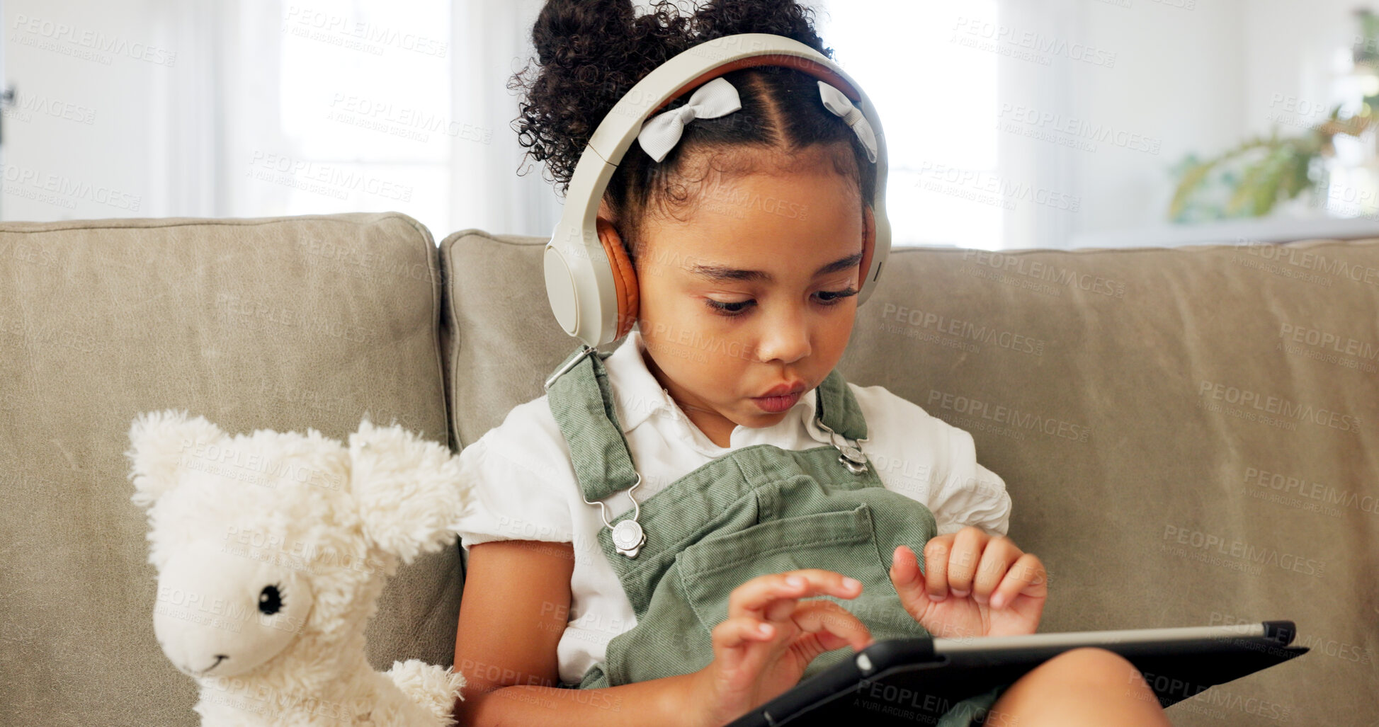 Buy stock photo Girl child, tablet and headphones on sofa, typing or reading for online course, thinking or education. Female kid, digital touchscreen and click for cartoon movie, video or gaming in home living room