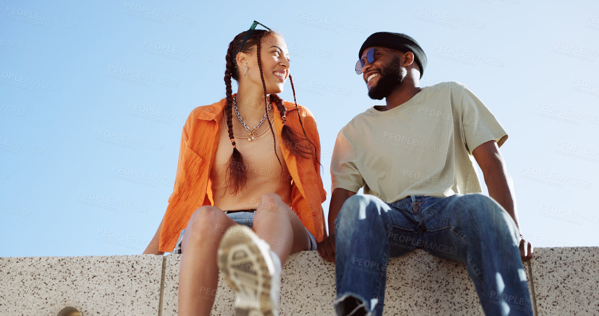 Buy stock photo Couple, city and smile with a black man, woman and fashion sitting on a wall, clear blue sky or bonding together. Street style, travel or love with a happy guy, girl or edgy with conversation outside