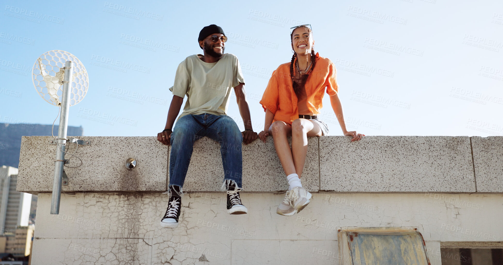 Buy stock photo Couple of friends, rooftop and date in city building, talking and youth in happy social bonding with summer sky. Diversity, gen z man and woman sit on urban roof with smile, love and relax together.