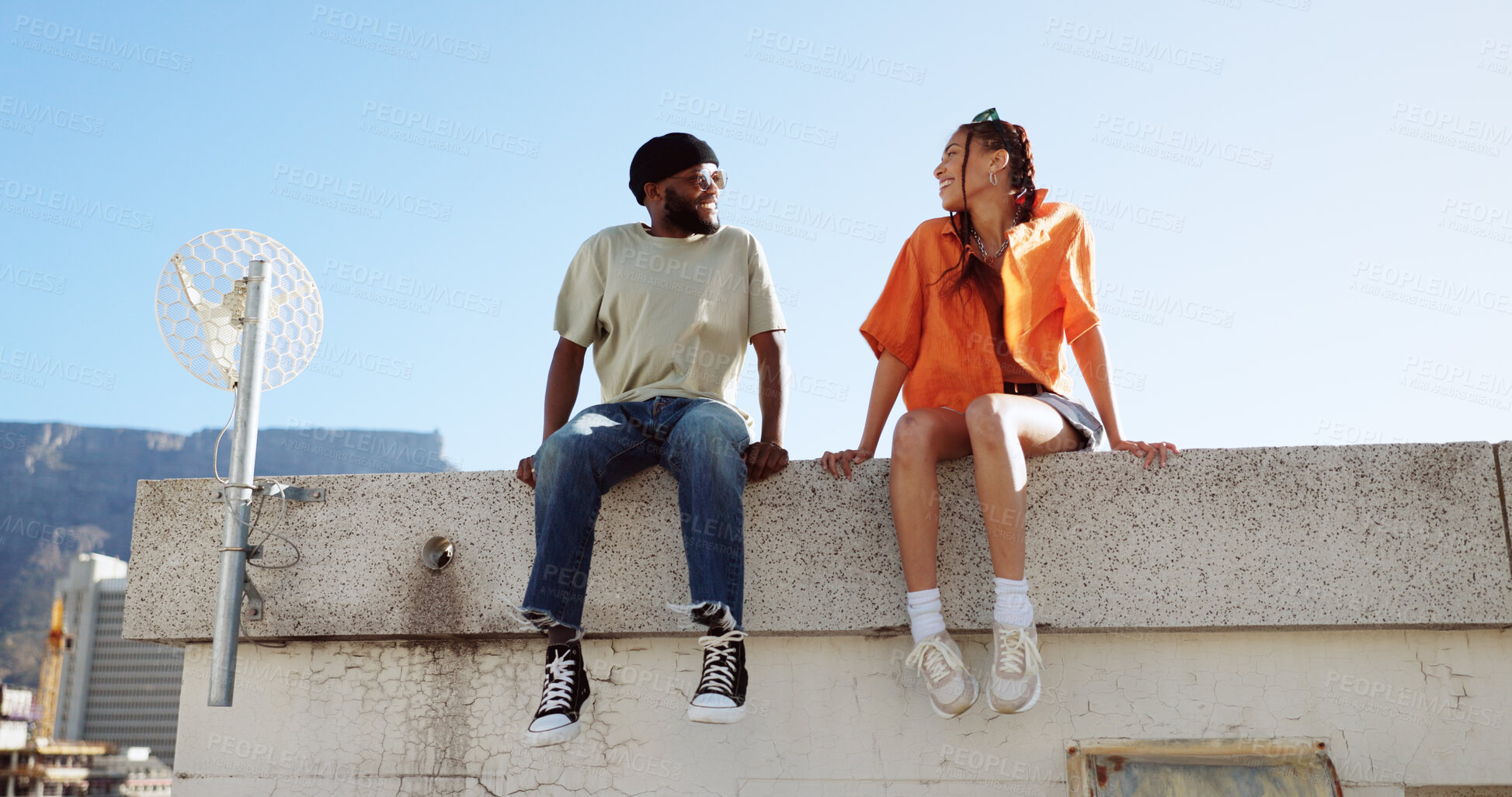Buy stock photo Couple of friends, rooftop and city building, talking and youth in happy social bonding with summer sky. Diversity, gen z man and woman sit on roof with smile, love and relax on urban date together.