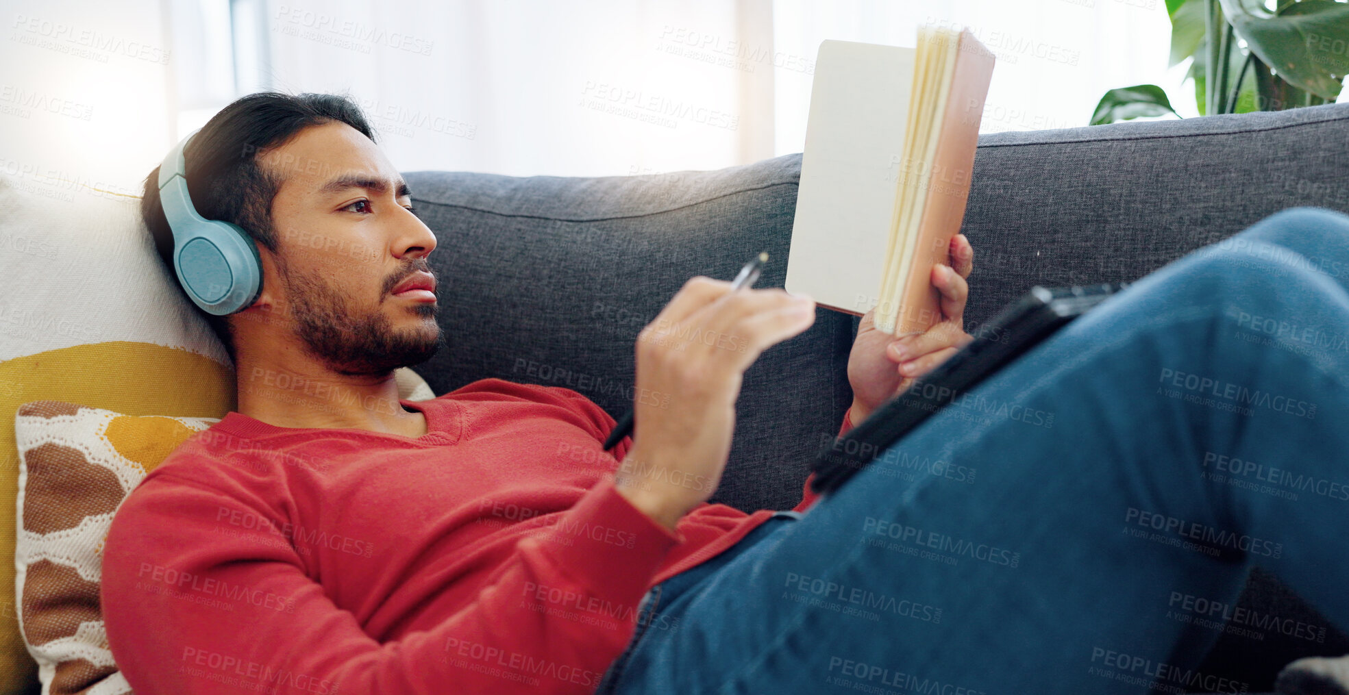 Buy stock photo Headphones, relax and man reading a book and listening to music, playlist or album on a sofa. Rest, technology and Indian male person enjoying a story and streaming a song in the living room at home.