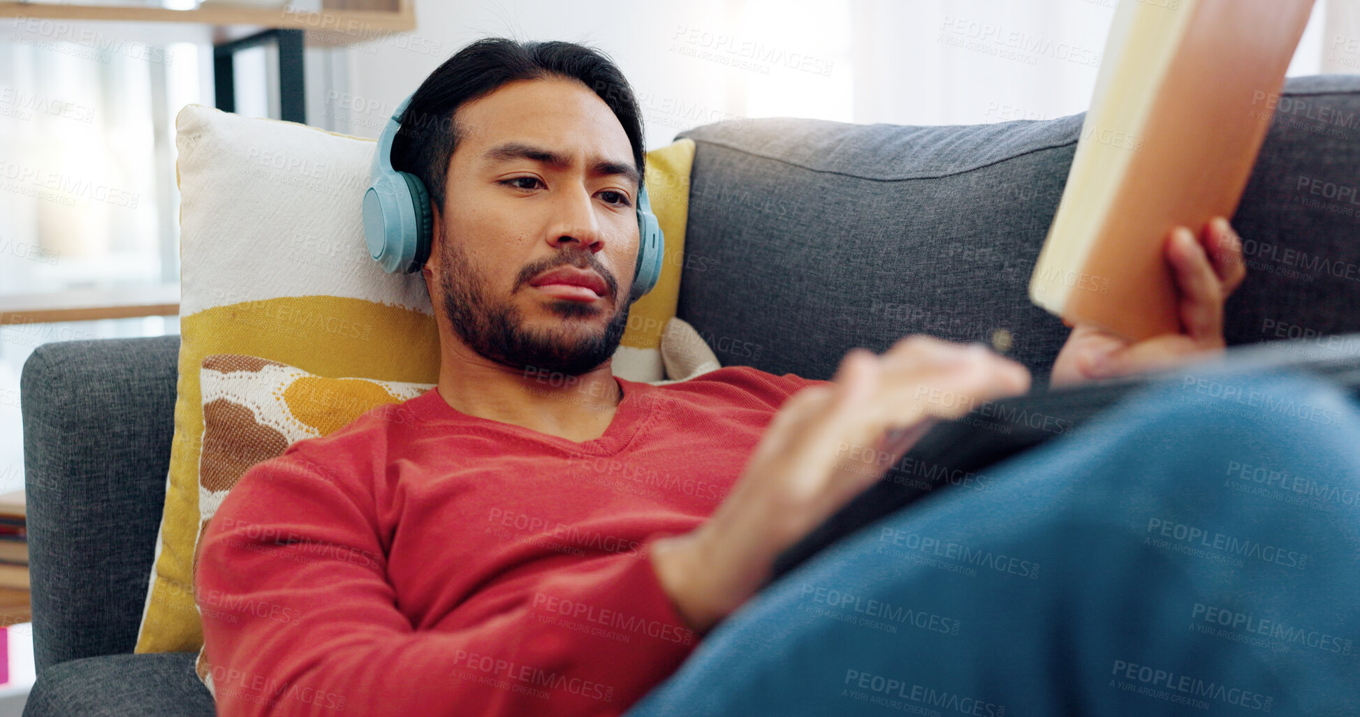 Buy stock photo Headphones, relax and man reading a story and listening to music, playlist or album on a sofa. Rest, technology and Indian male person enjoying a book and streaming a song in the living room at home.