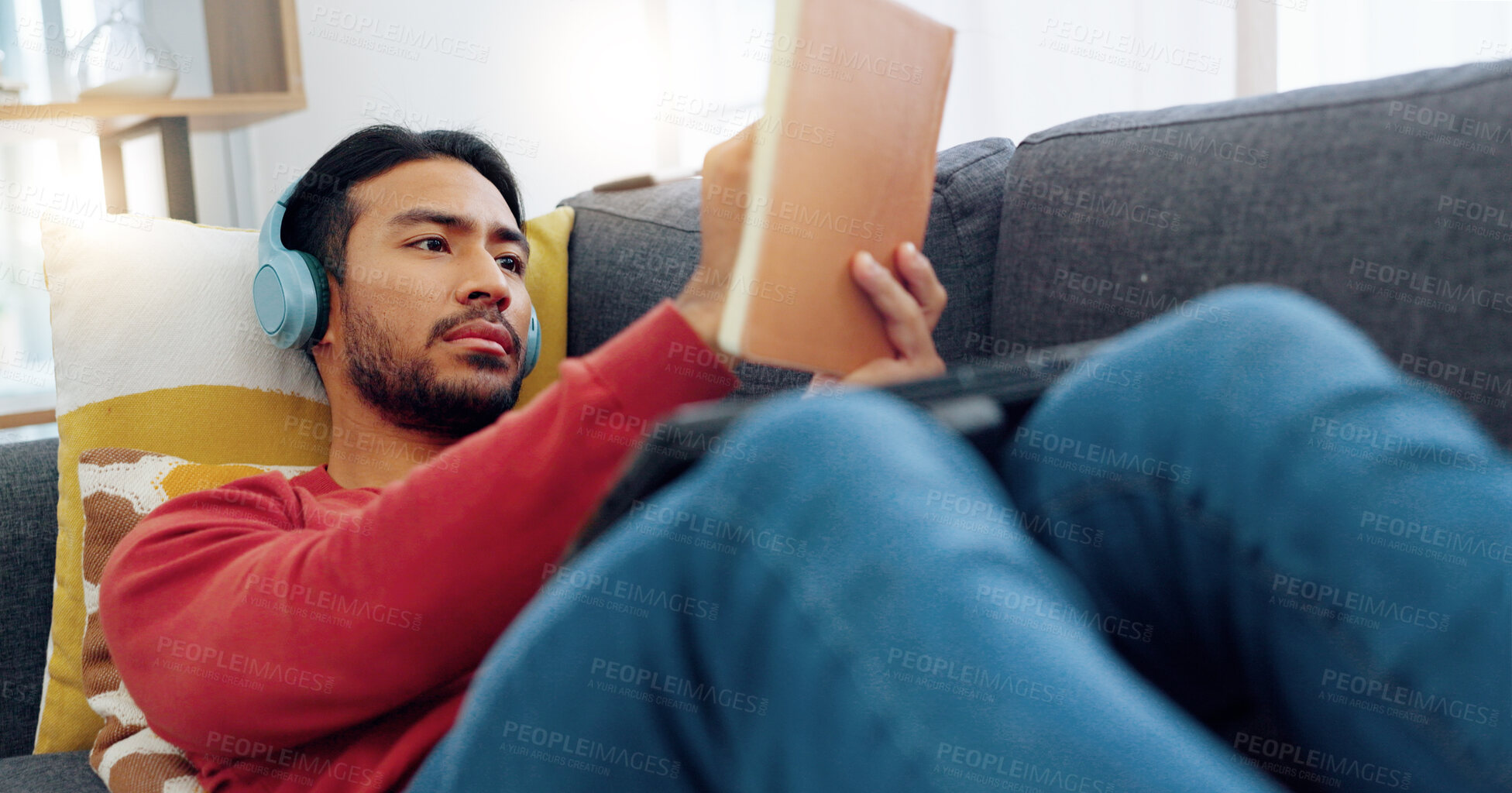 Buy stock photo Headphones, chilling and man reading a book and listening to music, playlist or album on a sofa. Relax, technology and Indian male person enjoying a story and streaming a song in living room at home.