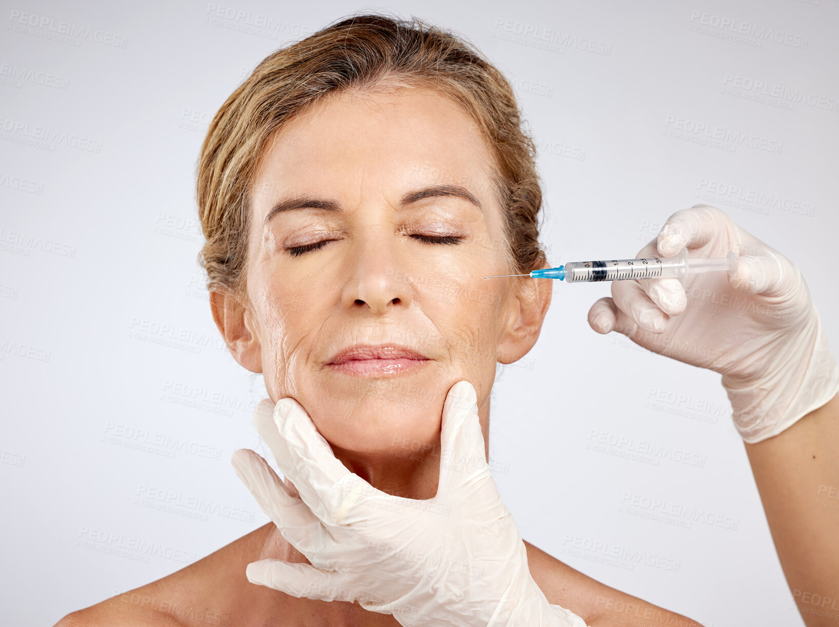 Buy stock photo Needle, surgery and face with mature woman getting an injection in her cheek for beauty, skincare and medicine in studio on a blue background. Filler, product and cosmetics with a female model inside