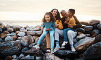Travel, beach and family sitting on rocks while on summer vacation, adventure or weekend trip. Happy, smile and portrait of mother bonding with her children and husband by the ocean while on holiday.