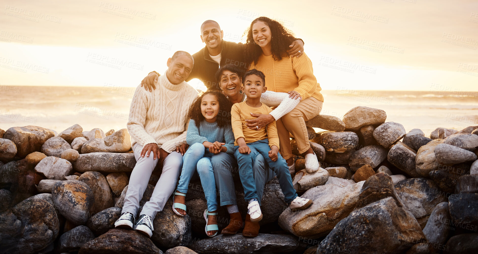 Buy stock photo Happy family, parents and children in beach portrait with excited face, sitting or relax with grandparents on rocks. Woman, man and kids by ocean for love, care and bonding on holiday by sunset sky