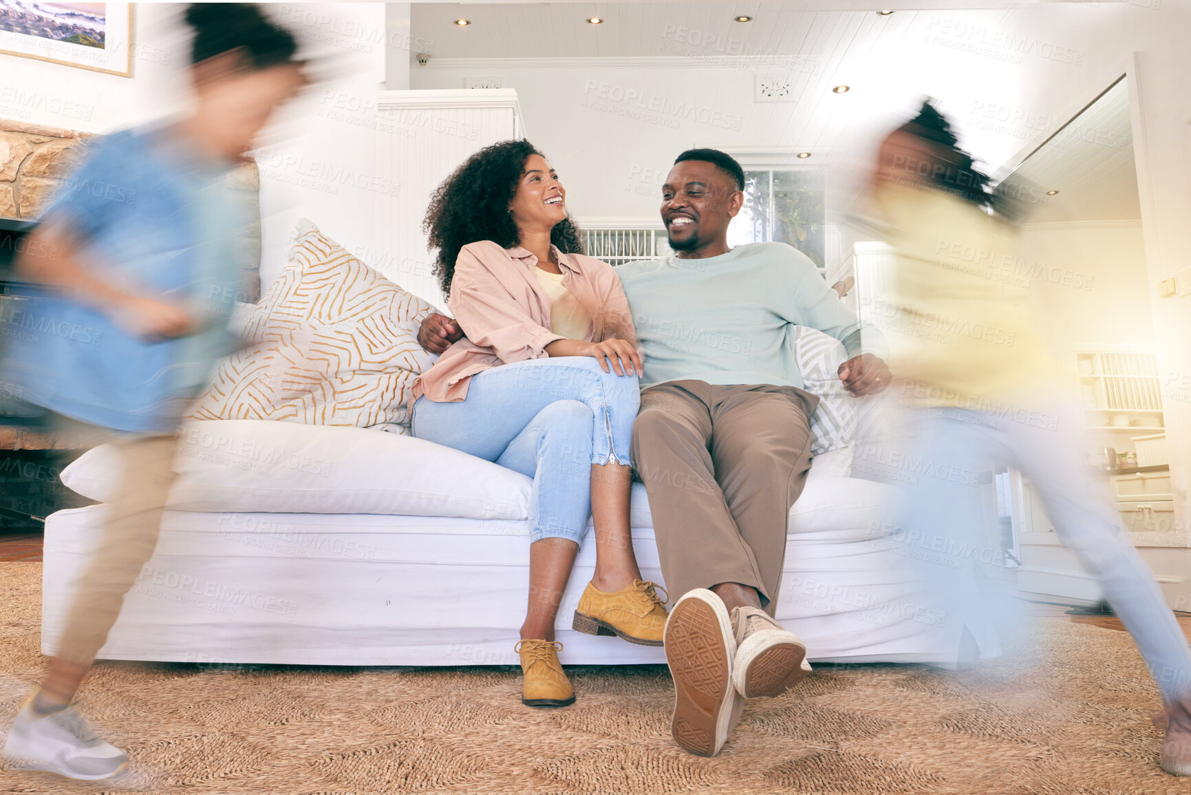 Buy stock photo Children running, happy home and couple on a sofa, talking and relax while bonding in a living room. Blur, kids and content parents on a couch, smile and enjoying weekend indoors with playing and fun