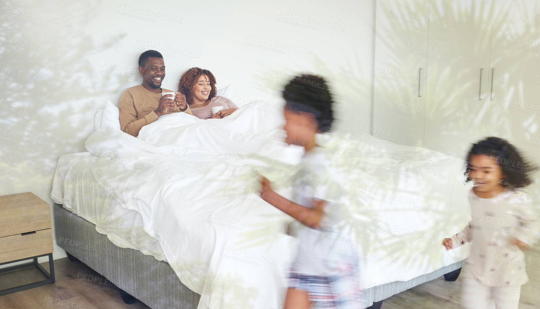 Buy stock photo Relax, family and children being playful in the bedroom while their parents lay in bed with a cup of coffee. Happy, love and young couple drinking a cappuccino while watching their kids run together.
